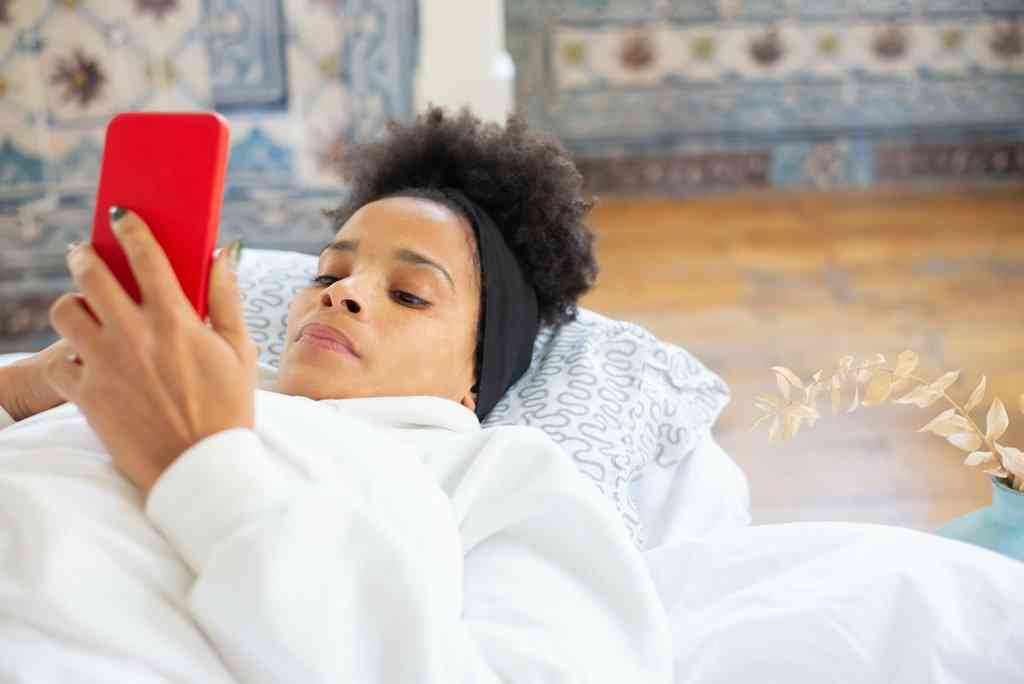 Женщина в белом халате лежит в кровати с красным смартфоном