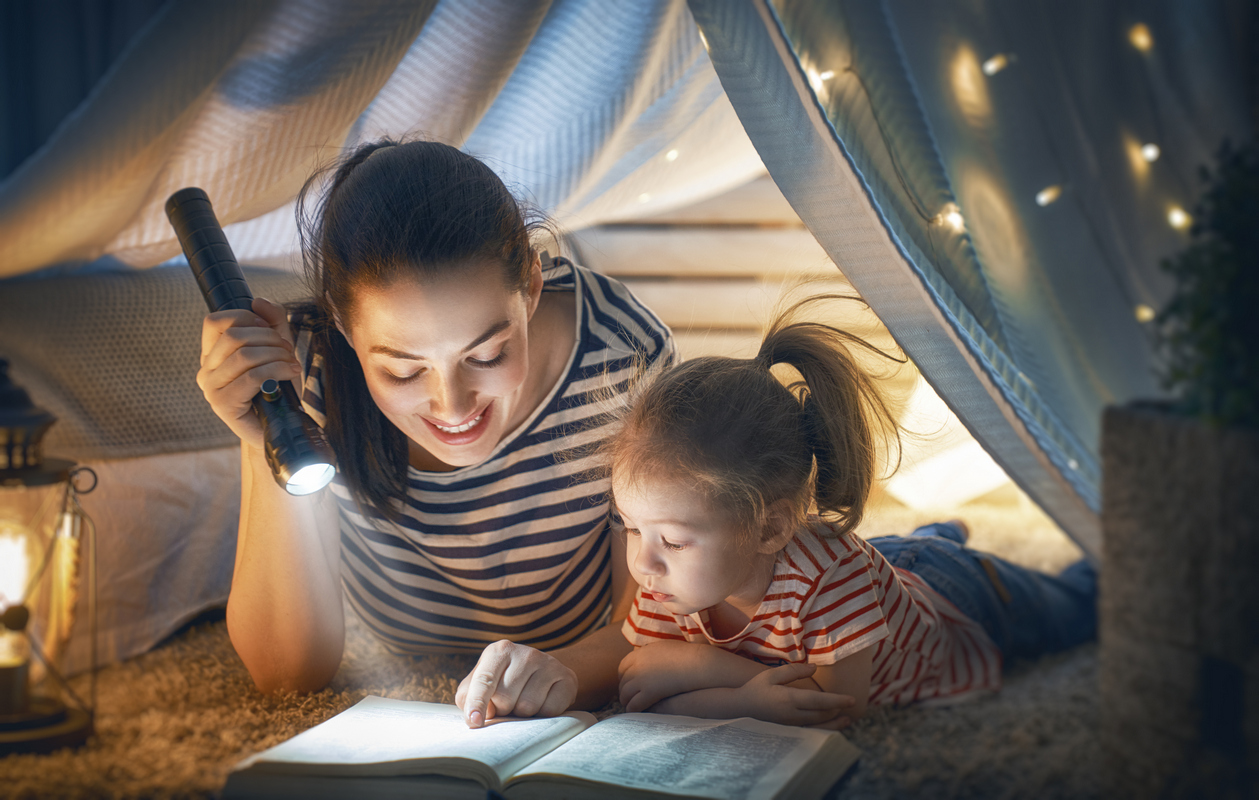 Мама с дочкой читают книжку с фонарем в шалаше